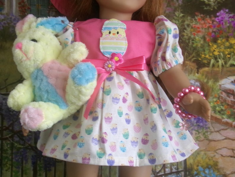 May include: A doll wearing a pink Easter dress with a white skirt patterned with Easter eggs. The doll holds a plush bunny in pastel colors. A pink bow and flower adorn the dress. A pink beaded bracelet is on the doll's arm.