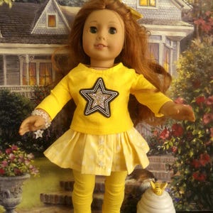 May include: A doll wearing a yellow outfit with a star design. The outfit includes a long-sleeved shirt, a skirt, and leggings. The doll has red hair and is wearing white shoes with yellow bows. A decorative beehive is next to the doll.