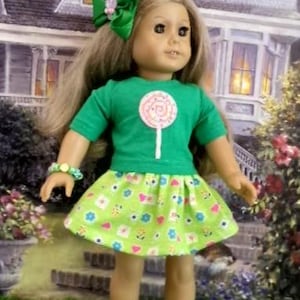 May include: A doll with long light brown hair wearing a green short-sleeved shirt with a white lollipop graphic, a light green skirt with a colorful floral and heart pattern, a green hair bow, a beaded bracelet, and light green clogs.