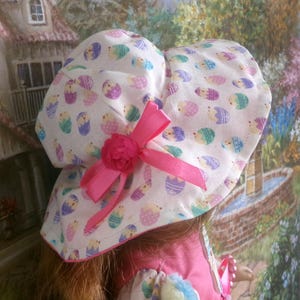 May include: A white Easter bonnet with a repeating pattern of colorful Easter eggs. The bonnet features a pink ribbon and a decorative pink flower. The brim is lined with pink fabric.
