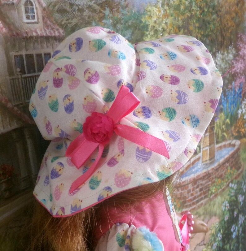 May include: A white Easter bonnet with a repeating pattern of colorful Easter eggs. The bonnet features a pink ribbon and a decorative pink flower. The brim is lined with pink fabric.