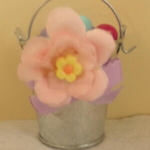 May include: A small, galvanized metal bucket with a silver handle, filled with a large pink fabric flower with a yellow center. The bucket is decorated with purple ribbon and contains colorful Easter eggs.