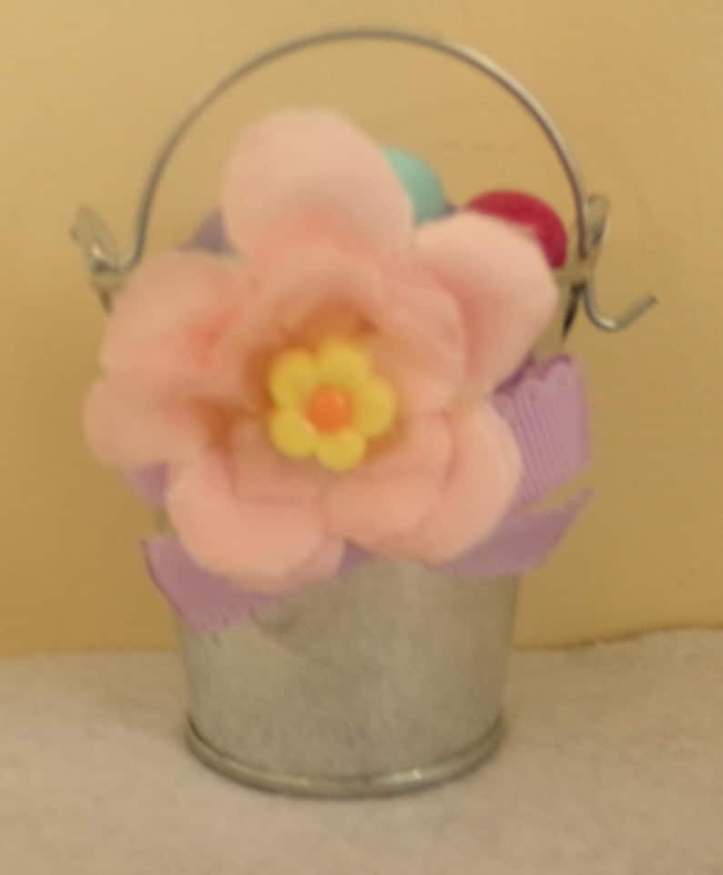 May include: A small, galvanized metal bucket with a silver handle, filled with a large pink fabric flower with a yellow center. The bucket is decorated with purple ribbon and contains colorful Easter eggs.