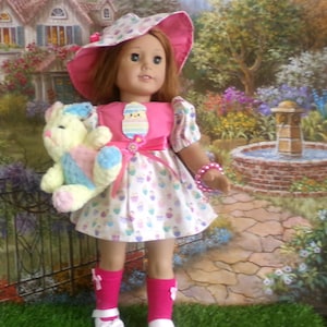May include: A doll wearing a pink dress with a white skirt patterned with cupcakes. The doll has a matching pink hat and holds a stuffed bunny. The doll is wearing pink socks and white shoes. A pink bracelet adorns the doll's wrist.