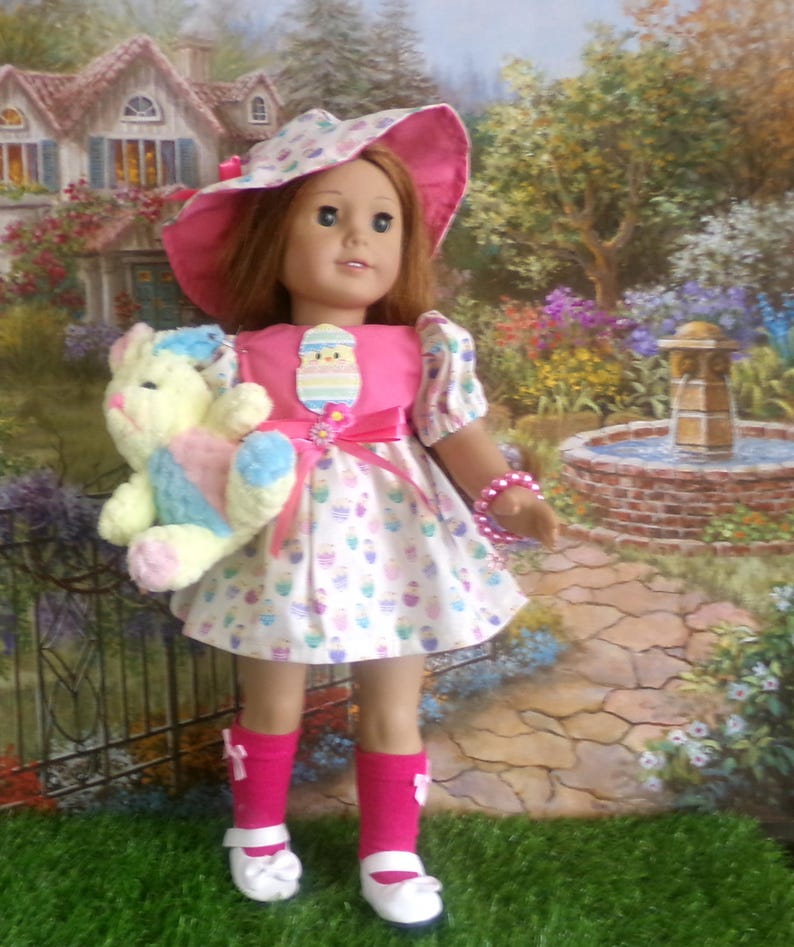 May include: A doll wearing a pink dress with a white skirt patterned with cupcakes. The doll has a matching pink hat and holds a stuffed bunny. The doll is wearing pink socks and white shoes. A pink bracelet adorns the doll's wrist.