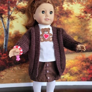 May include: A doll wearing a brown knitted cardigan, matching skirt, and white turtleneck. The cardigan has a teddy bear applique with a heart. The doll is also wearing white tights, black shoes, and a bracelet.