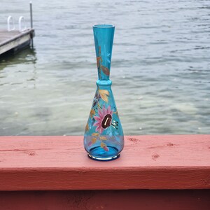 May include: A blue glass vase with a floral design painted on it. The vase has a narrow neck and a wide base. The flowers are painted in shades of pink, yellow, and green.