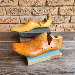 May include: Two antique wooden shoe forms, one complete and one in two pieces, sit on vintage books against a brick wall. The forms are a warm, aged wood colour, with the lower form displaying the text "470 ARMY".