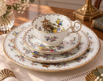 Könnte beinhalten: Ein weißes Porzellan-Teeservice mit Tasse und Untertasse, gestapelt auf einem passenden Speiseteller. Das Geschirr zeigt ein zartes Muster aus bunten Blumen, grünen Ranken und kleinen braunen Pagoden, verziert mit einem goldenen Rand.