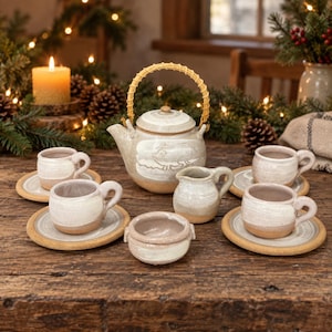 Puede incluir: Un juego de té de cerámica que incluye una tetera con asa trenzada, cuatro tazas con platillos, una pequeña jarra de leche y un azucarero. Cada pieza tiene un diseño bicolor con esmalte color crema y una base de tono natural.