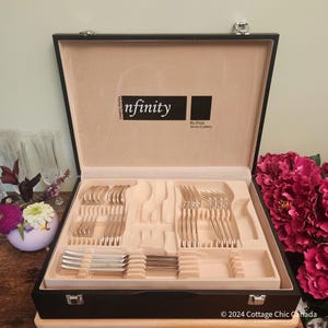 Könnte beinhalten: Ein schwarzer Besteckkoffer mit beigem Innenraum, der ein komplettes Silberbesteckset enthält. Der Koffer ist geöffnet und zeigt Gabeln, Löffel und Messer ordentlich angeordnet. Der Text "infinity" ist auf der Innenseite des Koffers aufgedruckt.