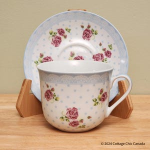 May include: A white teacup and saucer set with a floral rose pattern. The cup and saucer have a light blue border with a lace design and small blue polka dots. The roses are a deep pink color.