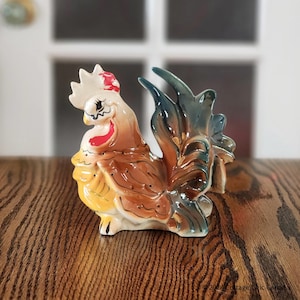 May include: A ceramic rooster ornament with a white crest and red details. The rooster features brown, yellow, and blue feathers. This decorative figurine is suitable for kitchen or farmhouse-style interiors.