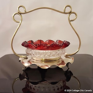 Può includere: Un portavivande in argento placcato con una ciotola in vetro rosso. Il portavivande ha una maniglia decorativa e tre piedini. La ciotola ha un bordo a festoni e una base in vetro trasparente.