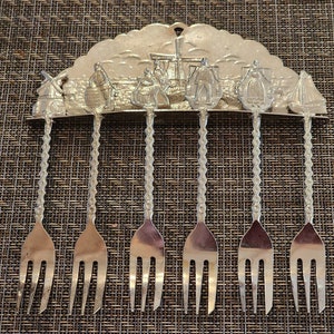 May include: Set of seven silver-toned forks with decorative windmill and Dutch scene handles. The forks are hanging on a silver-toned rack.