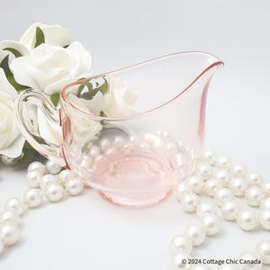 Puede incluir: Una jarra de crema o salsa de vidrio rosa con un asa transparente. La jarra está sobre una superficie blanca con una cadena de perlas blancas.