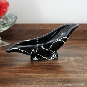 May include: A black ceramic ashtray with white abstract lines. The ashtray is shaped like a boat and has a smooth, glossy finish.