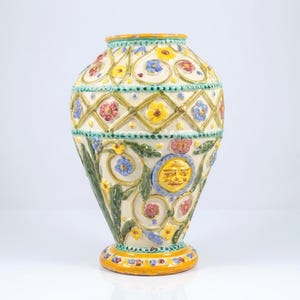 May include: A decorative ceramic vase with a cream base, decorated with colourful floral and geometric patterns. The vase features a sun-like face design and is accented with yellow, blue, and red hues. The vase is approximately 30 cm tall.