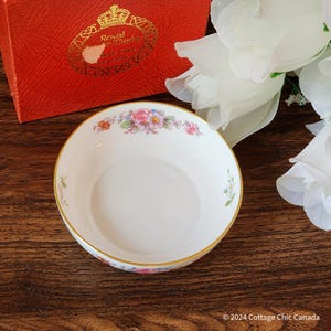 May include: A white porcelain bowl with a floral pattern in pink, purple, and yellow. The bowl has a gold rim. It is set on a wooden surface. A red box with gold lettering is in the background.