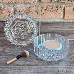 Puede incluir: Un tarro de vidrio transparente con tapa, la tapa tiene un patrón geométrico. El tarro contiene un compacto redondo y blanco de polvo prensado. También se muestra un pequeño pincel de maquillaje.