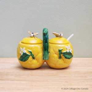 May include: A yellow ceramic sugar and creamer set shaped like lemons with green leaves and white flowers. The lids have small bee figurines on top.
