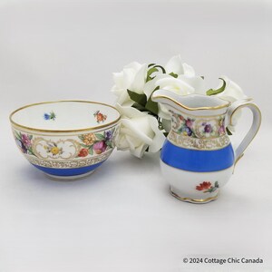 May include: A porcelain bowl and creamer set with floral and fruit designs. The bowl and creamer have a blue band and gold trim. The creamer has a handle and spout. White roses are in the background.