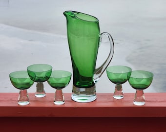 May include: A set of five green glass drinking glasses, including a pitcher, on a red wooden surface. The glasses are all the same shape and size, and the pitcher is taller than the glasses.