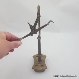 May include: Antique brass bottle opener with a hinged arm and a hook. The opener has a decorative base with a scalloped edge and several holes.
