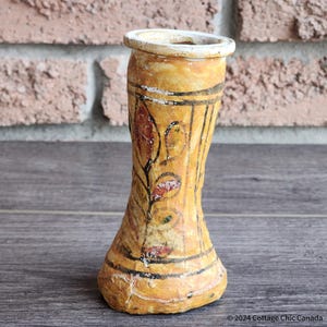Pode incluir: Um pequeno vaso de cerâmica amarelo com uma base alargada e um pescoço estreito. O vaso é decorado com desenhos florais pintados à mão em tons de vermelho, castanho e verde.