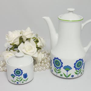 May include: White ceramic teapot and sugar bowl with a blue and green floral design. The teapot has a green rim and a spout. The sugar bowl has a lid.