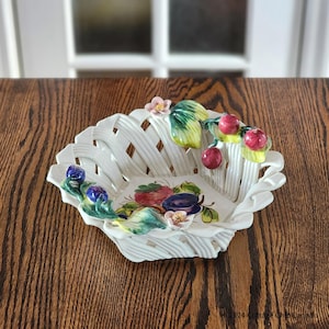 Puede incluir: Cesta de cerámica blanca con diseño tejido y borde festoneado. La cesta está decorada con coloridas frutas y flores de cerámica. La cesta está sobre una mesa de madera.
