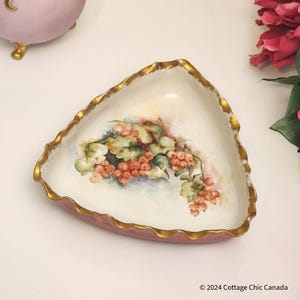 Puede incluir: Un plato triangular de estilo antiguo con un diseño pintado a mano de bayas rojas y hojas verdes. El plato tiene un borde festoneado con ribete dorado y una base rosa. La estética general es vintage y elegante.