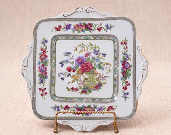 May include: A square white porcelain serving plate with scalloped edges and gold trim. The center features a colorful floral bouquet in a decorative vase, surrounded by a border of small flowers in shades of pink, purple, and green.