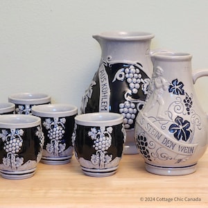 May include: A stoneware wine set with a pitcher and small cups. The set is mainly grey and black, with white grape cluster designs. The pitcher has a handle and a decorative relief of a figure. The cups are cylindrical.