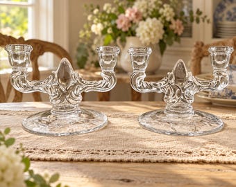 May include: A pair of clear glass candelabra featuring a central fleur-de-lis motif, two candle cups on curved arms, and a wide circular base with a starburst pattern. The glass is transparent with intricate molded details throughout.