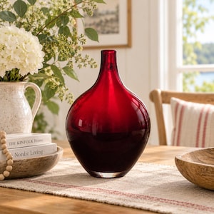 May include: A deep red glass vase with a rounded, bulbous body and a narrow, tapered neck. The glass is translucent, showing a smooth, polished surface that catches the light, creating a gradient of dark crimson tones throughout the vessel.