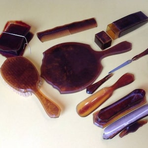 Puede incluir: Un juego de tocador vintage de carey con un cepillo, un peine, un espejo y varios recipientes. El juego está hecho de plástico marrón oscuro moteado.