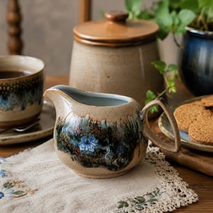 Peut inclure: Un pot à lait en céramique avec une base beige et une bande décorative de glaçure bleue, brune et noire. Le pot présente une anse incurvée, un bec verseur et un motif floral bleu peint à la main centré sur le devant du récipient.