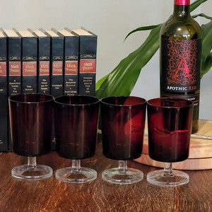 Könnte beinhalten: Vier rote Weingläser mit klaren Stielen und Füßen stehen auf einem Holztisch neben einer Flasche Apothic Red Wein und einem Stapel Bücher mit dem Namen "Mark Twain" auf den Rückseiten.
