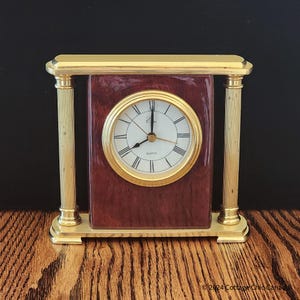 May include: A rectangular quartz clock with a gold-coloured frame and two gold columns. The clock face is white with black Roman numerals and gold hands. The clock's body is a dark reddish-brown colour.
