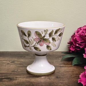 Op de afbeelding: Witte keramische voetbalkom met een bloemmotief met roze en groene accenten. De kom heeft een brede, open bovenkant en een decoratieve basis met gouden rand. Het bloemmotief bevat een vlinder en bladeren.