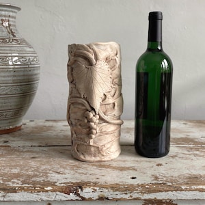 May include: A beige, cylindrical vase with a grape leaf and vine design. A green wine bottle with a black cap is next to the vase. A ceramic vase is in the background. The objects are on a distressed wooden surface.