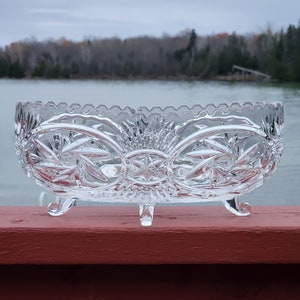 May include: A clear glass bowl with a cut crystal design and three legs. The bowl is oval shaped and has a scalloped edge.