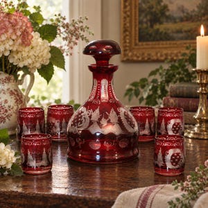 May include: A ruby red glass decanter with a matching stopper and five coordinating glasses, all featuring intricate etched floral and scroll patterns. The set is crafted from deep red glass with clear cut-glass details throughout the design.