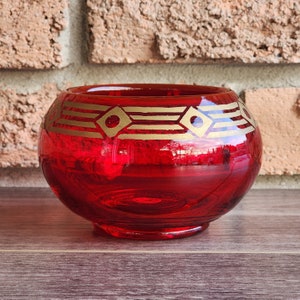 Peut inclure: Un bougeoir en verre rouge avec un motif géométrique doré. Le bougeoir a une forme arrondie et une large ouverture.