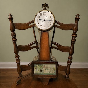 May include: A wooden wall clock featuring a round face with black Arabic numerals and a brass eagle finial at the top. The clock body includes a rectangular panel at the base displaying a painted landscape scene of a large building with white columns.