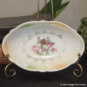 Puede incluir: Un plato ovalado blanco para servir con un borde festoneado y un diseño floral. El plato tiene un borde dorado y un delicado patrón floral con rosas rosas y blancas.
