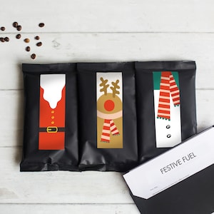 May include: Three black coffee bags with Christmas-themed designs: Santa Claus, a reindeer, and a snowman. The Santa bag has a red suit and gold belt. The reindeer has antlers and a scarf. The snowman has a red and white scarf. Coffee beans are scattered.