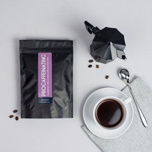 May include: A black bag of decaffeinated coffee with the text "PROCAFFEINATING to postpone or delay action until you had coffee" and the brand name "NOVELLO" printed on the front. A black coffee pot, a white cup of coffee, and a silver spoon are on a white surface.