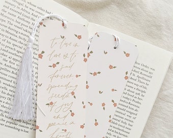 May include: Two floral bookmarks with white tassels rest on an open book. One bookmark features a quote: "To live in love is to say forever, spreading seeds of joy and peace in hearts." The other has a floral pattern.